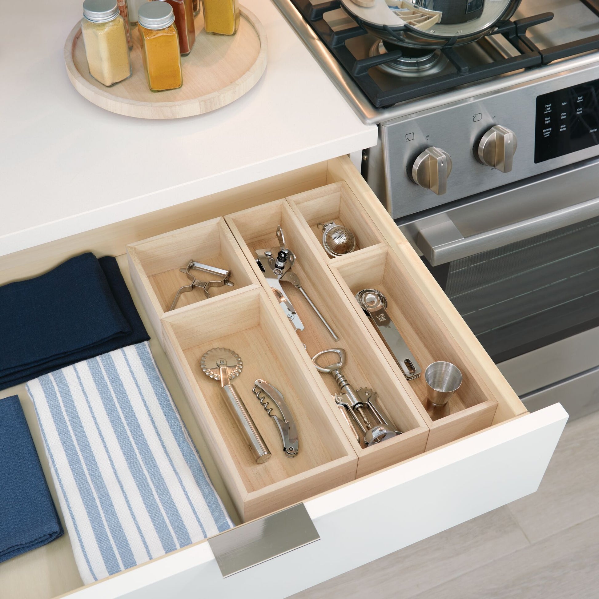 A light natural wood drawer organizer insert with multiple compartments, shown installed in an open kitchen drawer containing metal kitchen tools. The scene includes a white drawer front, a stainless steel gas stove, folded blue towels, and spice jars on a countertop.