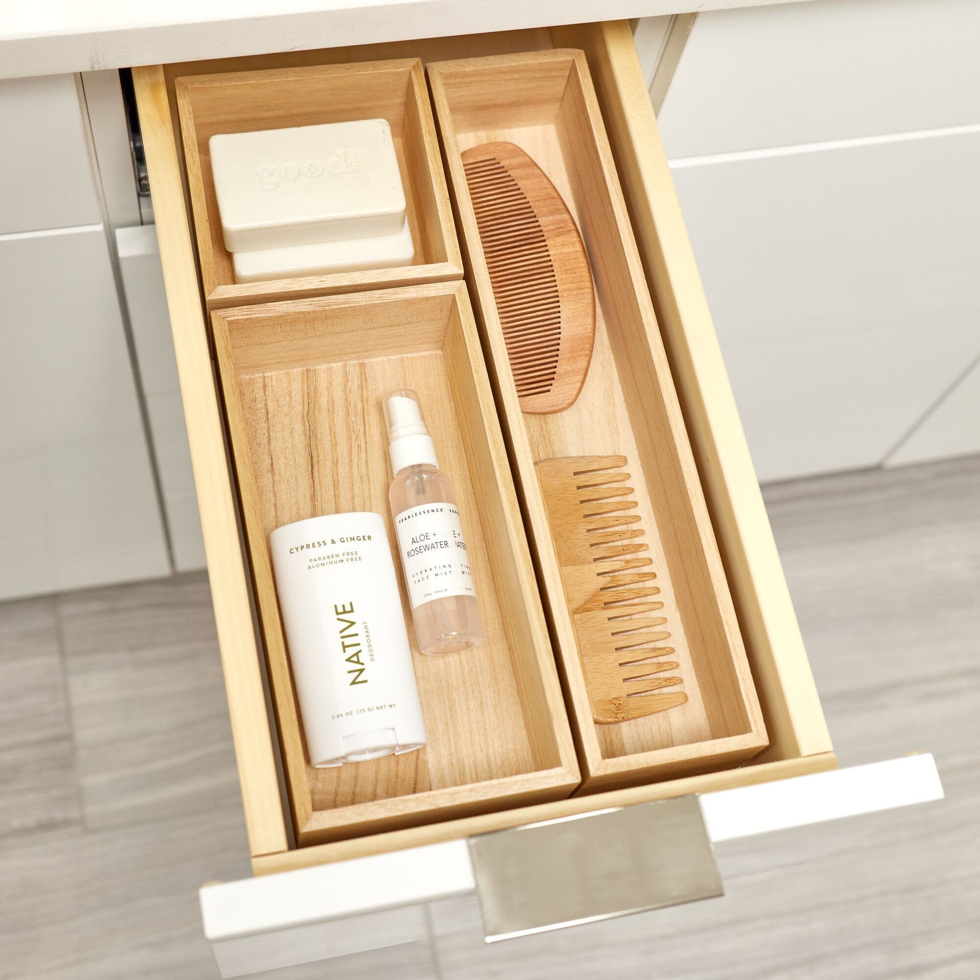 A wooden drawer organizer set arranged inside an open drawer, holding a white soap bar, a small spray bottle, and two wooden hair/combing tools. The scene is shown in a bathroom drawer with cabinetry and floor visible.