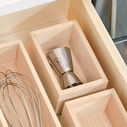 A stainless steel jigger with a compass logo, nestled inside a light natural wood drawer organizer tray.