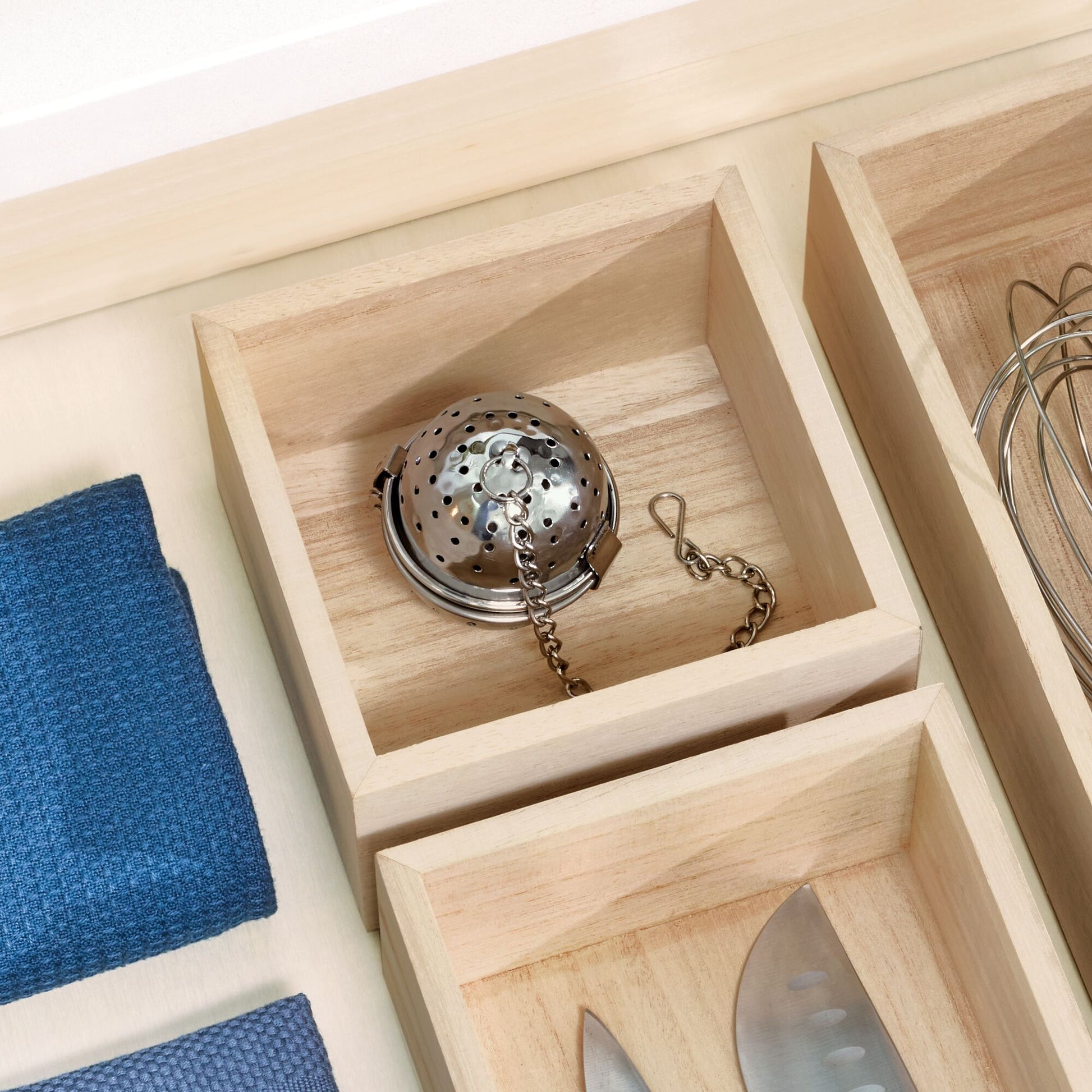 A close-up of a light natural wood drawer organizer with multiple compartments. One compartment holds a stainless steel tea infuser, while another holds a metal whisk.