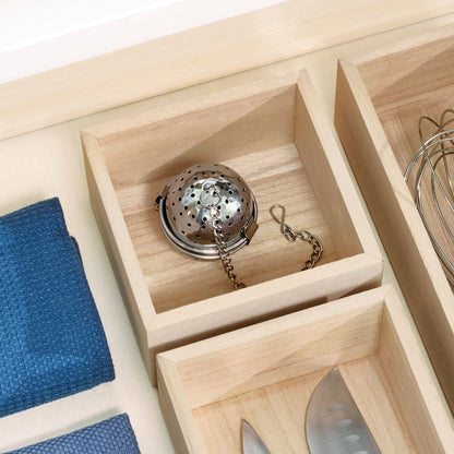 A close-up of a light natural wood drawer organizer with multiple compartments. One compartment holds a stainless steel tea infuser, while another holds a metal whisk.
