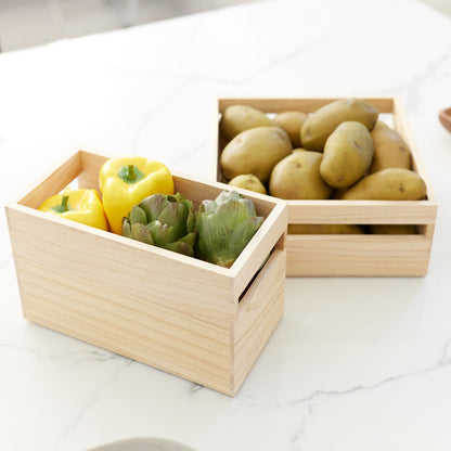 Two light natural wood storage boxes on a white marble countertop, one filled with yellow bell peppers and green artichokes, and the other with potatoes.